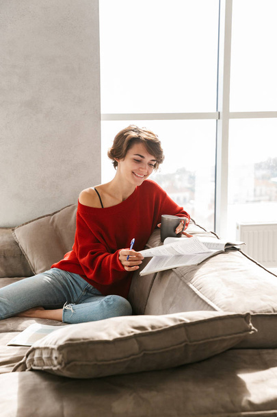 Cheerful girl studying at home while laying on a couch and drinking tea - Photo, image