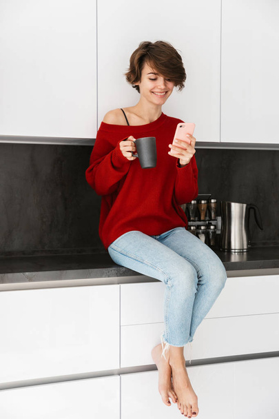 Smiling younh woman wearing sweater sitting on a kitchen table at home, using mobile phone, drinking coffee - Fotoğraf, Görsel