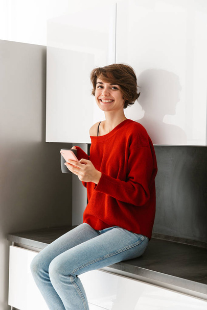 Smiling younh woman wearing sweater sitting on a kitchen table at home, using mobile phone, drinking coffee - Foto, imagen