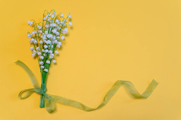 Boeket van lelies van de vallei bloemen met groen lint op fel geel papier achtergrond. Mooie lente samenstelling in minimalistische stijl. Bovenaanzicht, vlakke lay, kopieer ruimte - Foto, afbeelding