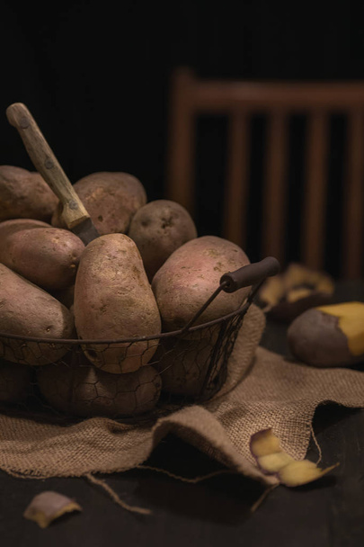 dunkle und stimmungsvolle, mystische leichte Food-Fotografie. Kartoffeln, die mit einem Schälmesser auf Holzgrund geschält werden. rustikales Stillleben mit natürlicher, gerichteter Beleuchtung zur Betonung von Textur und Details.  - Foto, Bild