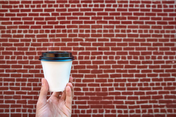 Close up of male hand holding white paper cup of coffee, against a red brick wall. Enjoying coffee to go. Fast food. Business or food and drink concept. Space for text - Photo, image