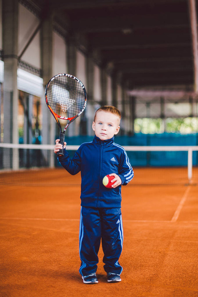 Σεπτεμβρίου 20, 2016. Ουκρανία, Kiev.theme σωματική ανάπτυξη παιδιών. Καυκάσιος παιδί αγόρι 3 - 4 χρόνια παλιό πόζες με τις ρακέτες τένις τένις tnet φόντο πορτοκαλί φλοιό χέρι στο μπλε κοστούμι αθλητισμού. - Φωτογραφία, εικόνα