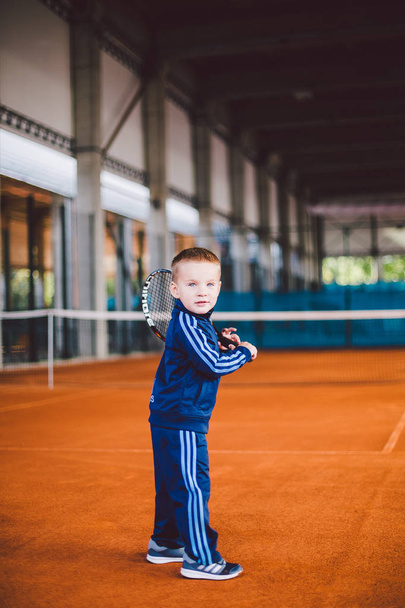 20 de setembro de 2016. Ucrânia, Kiev.theme crianças desenvolvimento físico. Menino caucasiano de 3 a 4 anos posa com raquete de tênis bola mão tênis laranja casca de fundo tnet em azul terno esportivo
. - Foto, Imagem
