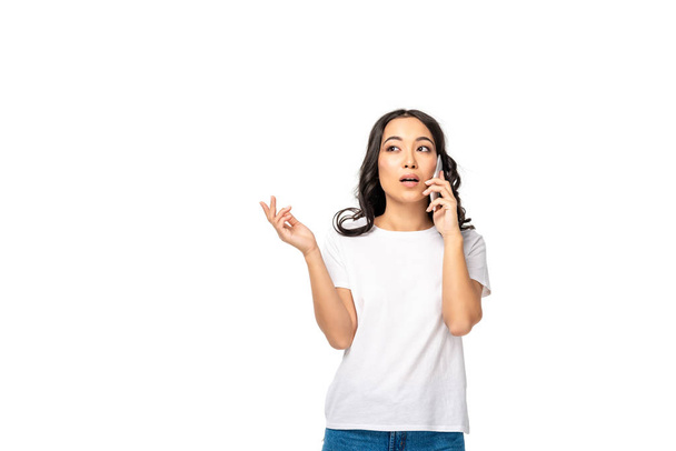 Thoughtful asian girl in white t-shirt and blue jeans talking on smartphone isolated on white - Foto, immagini