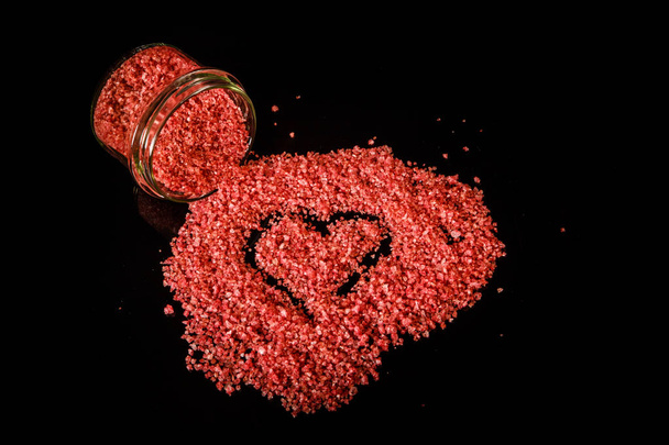 Χειροποίητη καρδιά σε ροζ κόκκινο χρώμα φυσικό οργανικό αλάτι χύνεται έξω από το μικρό γυάλινο βάζο σε φόντο μαύρο καθρέφτη - Φωτογραφία, εικόνα
