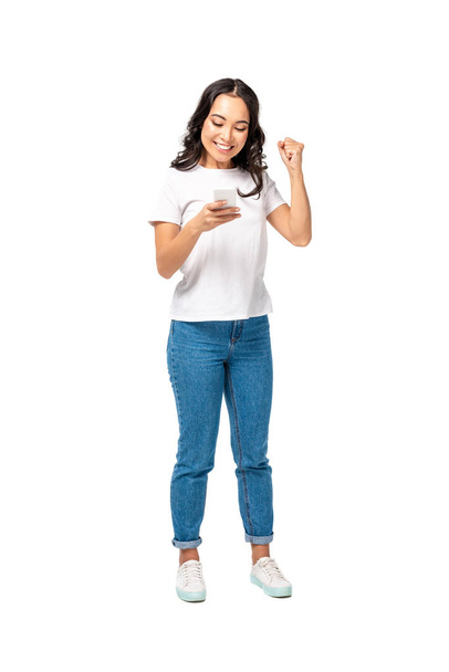 Smiling happy asian girl in white t-shirt and blue jeans using smartphone and raising fist isolated on white - Photo, Image