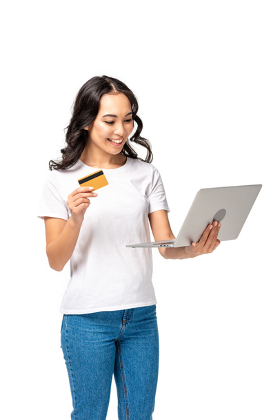 Hymyilevä kaunis aasialainen nainen valkoisessa t-paidassa ja sinisissä farkuissa kannettavan tietokoneen avulla ja pitämällä luottokorttia eristettynä valkoisella
 - Valokuva, kuva