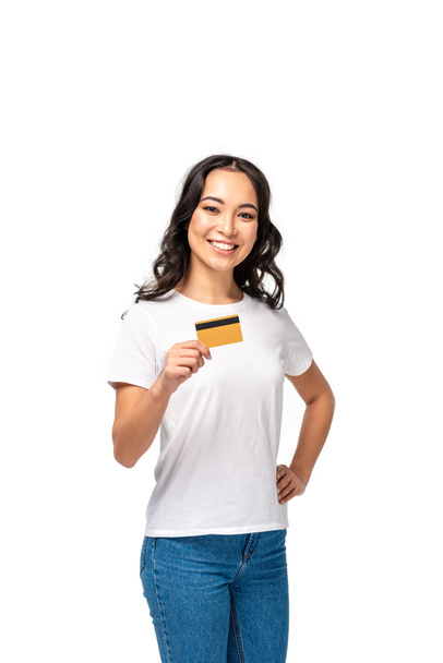 Beautiful smiling asian woman showing credit card and holding hand on hip isolated on white - Foto, afbeelding