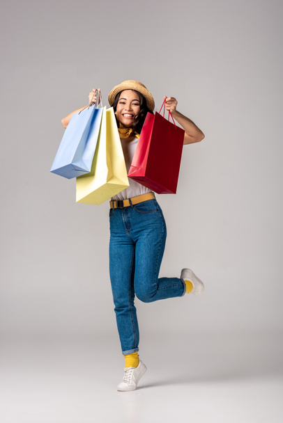 Happy trendy dressed asian woman holding colorful shopping bags on raised hand on grey - Zdjęcie, obraz