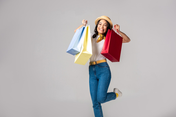 Happy trendy dressed asian woman holding colorful shopping bags on raised hands isolated on grey - Fotó, kép