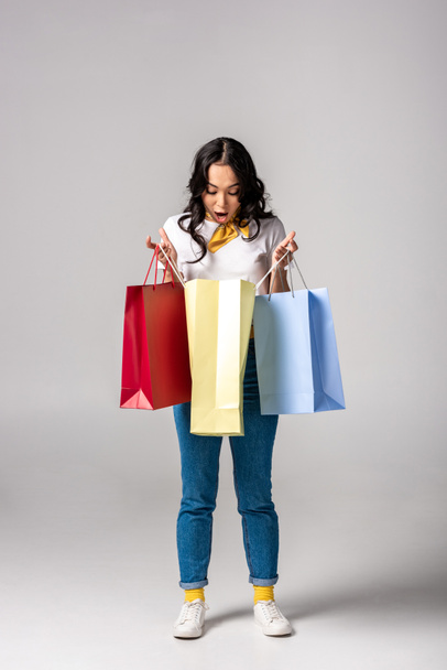 Surprised young asian woman looking inside colorful shopping bags on grey - 写真・画像