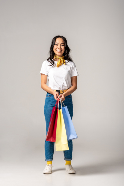 attraktive lächelnde asiatische Frau mit bunten Einkaufstaschen auf grau - Foto, Bild