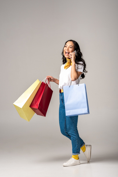 Sonriente joven asiática mujer sosteniendo coloridas bolsas de compras y hablando en el teléfono inteligente en gris
 - Foto, Imagen