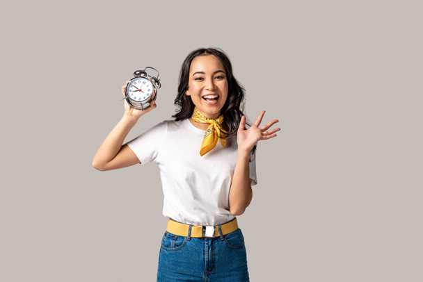 Счастливая азиатка в белой футболке и синих джинсах держит будильник и машет рукой, изолированной на сером
 - Фото, изображение