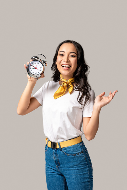 smiling young asian woman in white t-shirt and blue jeans holding alarm clock isolated on grey - Photo, Image