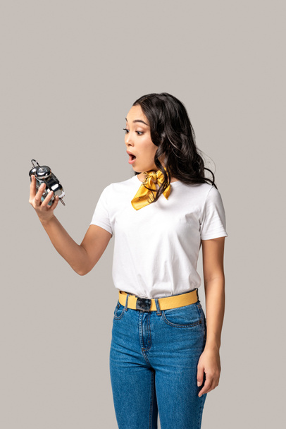 Surprised young asian woman in white t-shirt and blue jeans holding alarm clock isolated in grey  - Photo, Image