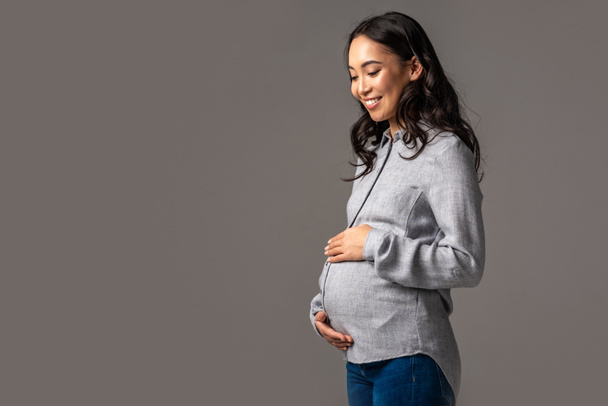 Smiling pregnant asian woman holding tummy with hands isolated on grey - Foto, Imagem