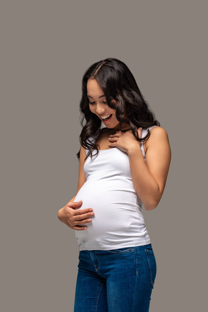 Χαμογελώντας έγκυος γυναίκα της Ασίας σε λευκό t-shirt και τζιν μπλε κοιτάζοντας κοιλιά που απομονώνονται σε γκρι - Φωτογραφία, εικόνα