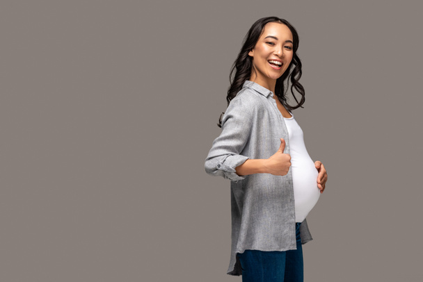 lächelnd schwangere Asiatin berührt Bauch und zeigt Daumen nach oben isoliert auf grau - Foto, Bild