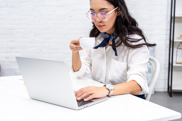 Young asian freelancer in glasses using laptop and drinking coffee at home workplace - Zdjęcie, obraz