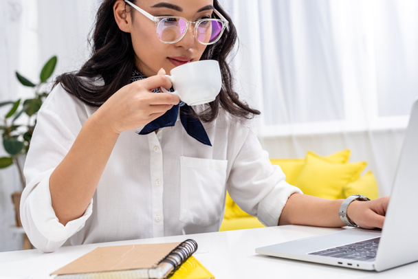 Atraktivní asijské freelancer pomocí přenosného počítače a pití kávy na doma pracovišti - Fotografie, Obrázek