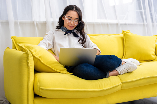 Bastante sonriente freelancer asiático usando portátil mientras está sentado con los pies en el sofá amarillo en casa
 - Foto, Imagen