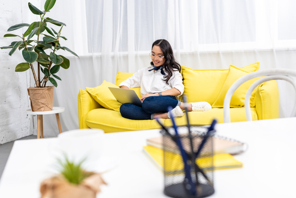 selektiver Fokus asiatischer Freelancer, die mit Füßen auf gelbem Sofa sitzen und Laptop mit verschwommenem Arbeitsplatz im Vordergrund benutzen - Foto, Bild