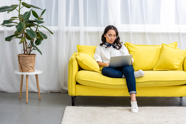 Átgondolt ázsiai szabadúszó szemüveget használ a laptop otthon sárga kanapén kényelmesen ülve - Fotó, kép