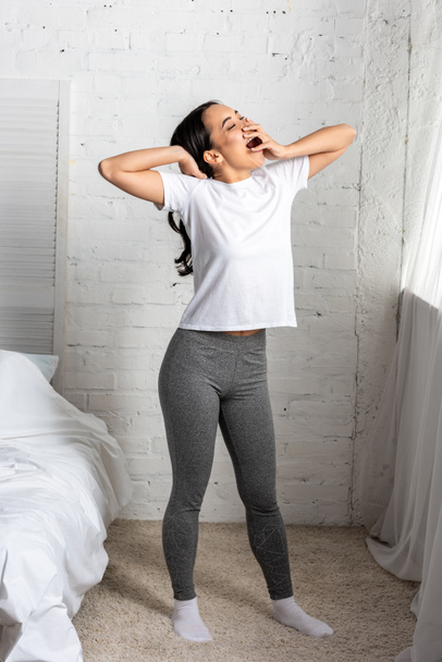 Young asian woman in white t-shirt and grey leggings standing in bedroom in yawning - Photo, Image