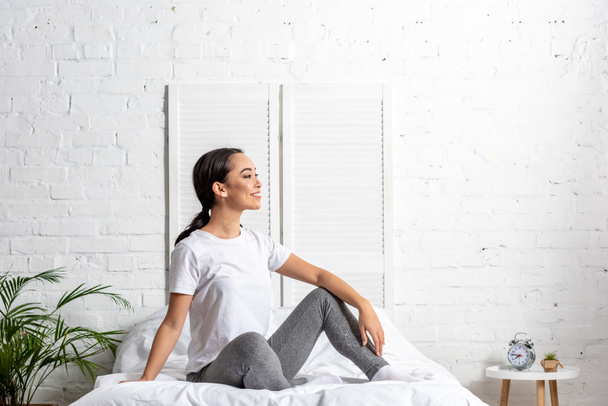 mujer asiática en camiseta blanca y polainas grises sentada en la cama y descansando después de hacer ejercicio por la mañana
 - Foto, Imagen