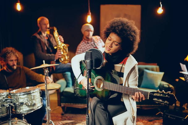 Mixed race woman singing and playing guitar while sitting on chair with legs crossed. In background drummer, saxophonist and bass guitarist. - Photo, Image