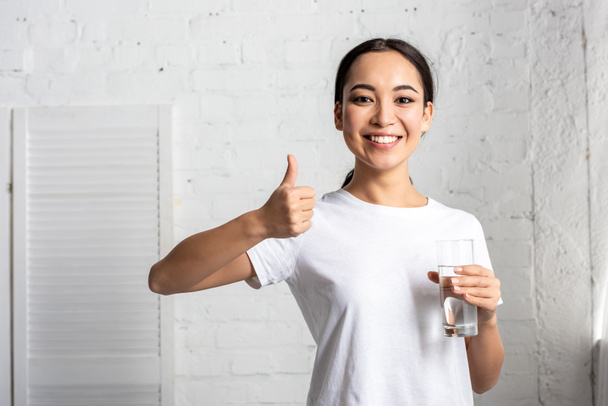 lachende jonge Aziatische vrouw in wit t-shirt glas water te houden en het tonen van de duim omhoog - Foto, afbeelding