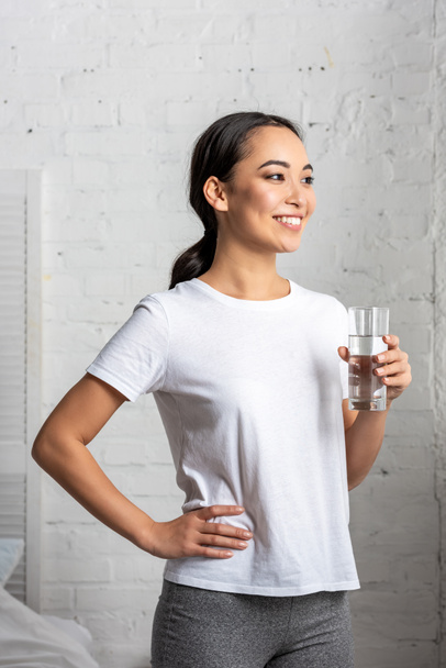 smiling young asian  woman holding glass of water, standing in bedroom with hand on hip - Foto, Imagem