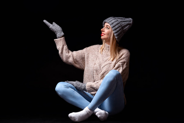 Una chica rubia sonriente en guantes de punto gris, sombrero y suéter sentado en el suelo negro con las piernas cruzadas con los ojos hacia arriba y coger copos de nieve por el brazo sostenido, sobre fondo negro aislado
 - Foto, imagen