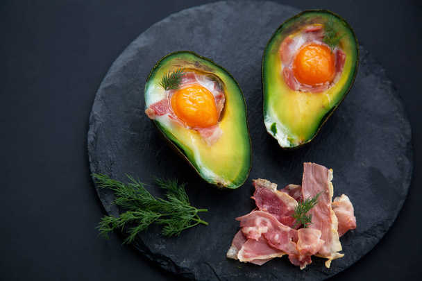 two baked avocado halves with egg and sliced becon on black stone Board,close up - Photo, Image