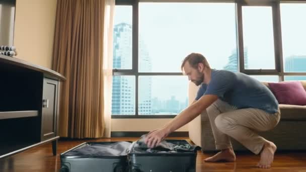 ハンサムな男が高層ビルを見渡すパノラマ ビューの窓と部屋でスーツケースをパックします。 - 映像、動画