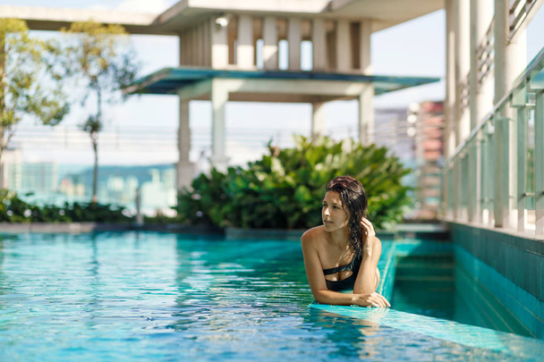 Retrato de uma mulher caucasiana sorridente sexy em um maiô estava na borda de uma piscina no último piso com arbustos verdes e vistas da cidade. Ásia fim de semana
. - Foto, Imagem