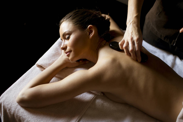 Mulher bonita tendo seu ombro e corpo massageados com pedras quentes por um massagista profissional em um centro de spa
 - Foto, Imagem