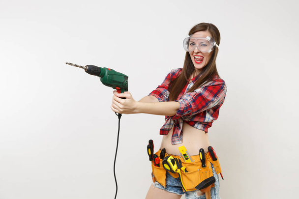 Strong young energy handyman woman in plaid top shirt denim shorts kit tools belt full of instruments in protective goggles hold power electric drill isolated on white background. Female in male work - Photo, Image