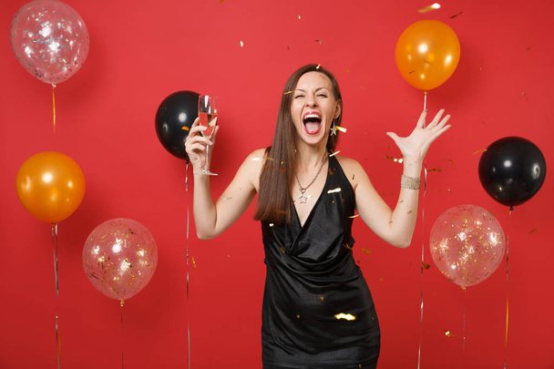 Mujer joven loca en vestido negro gritando celebrando, sosteniendo una copa de champán y atrapando confeti en globos de aire de fondo rojo brillante. Feliz Año Nuevo, concepto de fiesta de cumpleaños simulada
 - Foto, Imagen