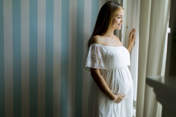 Kaunis nuori raskaana oleva nainen seisoo ikkunan vieressä
 - Valokuva, kuva