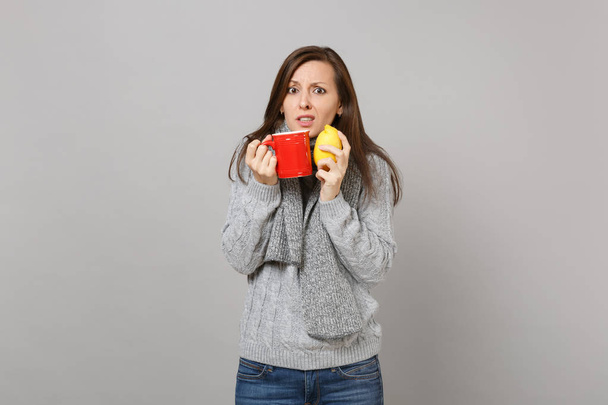 Puzzled young woman in gray sweater, scarf holding lemon, red cup of tea isolated on grey wall background. Healthy fashion lifestyle, people sincere emotions, cold season concept. Mock up copy space - Fotografie, Obrázek