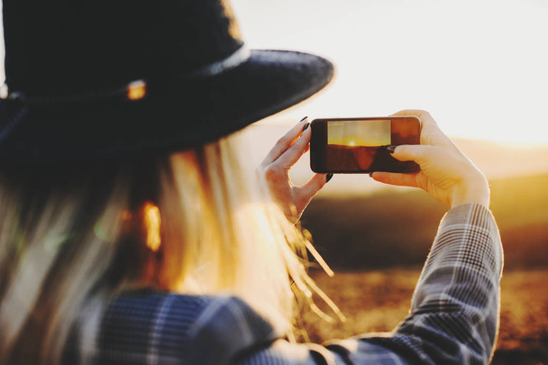 Back view of young female using smartphone to take pictures of setting sun in wonderful countryside.Anonymous woman taking pictures of sunset - Photo, Image