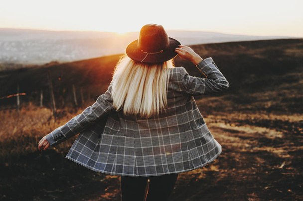 Visão traseira da jovem fêmea em chapéu da moda e jaqueta em pé no campo incrível durante o pôr do sol magnífico.Mulher anônima no campo durante o pôr do sol
 - Foto, Imagem