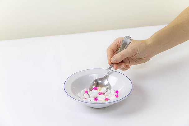 Пристрастие к лекарствам и наркотикам в наше время: рука молодой женщины с ложкой таблеток, взятых из тарелки, полной лекарств на белом столе и фоне
 - Фото, изображение