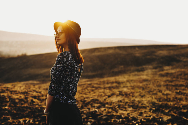 Vista lateral de la bella dama joven en traje elegante mirando hacia otro lado mientras está de pie en el campo de otoño durante la maravillosa puesta del sol.
 - Foto, Imagen