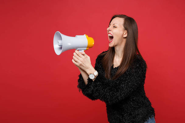 Portrait of crazy young girl in black fur sweater looking aside screaming on megaphone isolated on bright red wall background in studio. People sincere emotions, lifestyle concept. Mock up copy space - Photo, Image