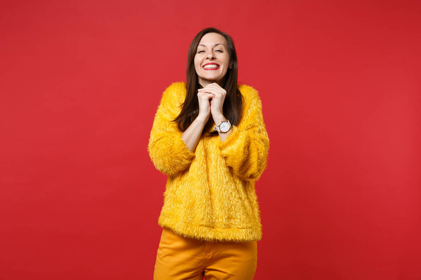 Portrait d'heureuse jolie jeune femme en pull en fourrure jaune mettant les mains près du menton isolé sur fond de mur rouge vif en studio. Les gens émotions sincères, concept de style de vie. Maquette espace de copie
 - Photo, image
