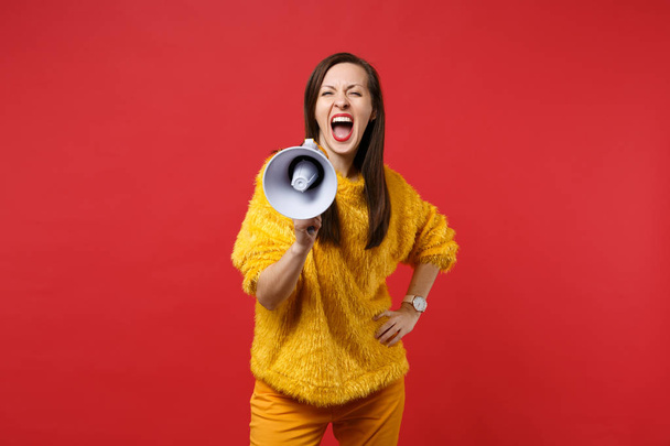 Retrato de mujer joven loca en suéter de piel amarilla de pie, gritando en megáfono aislado en fondo de pared de color rojo brillante en el estudio. Gente emociones sinceras, concepto de estilo de vida. Simular espacio de copia
 - Foto, Imagen